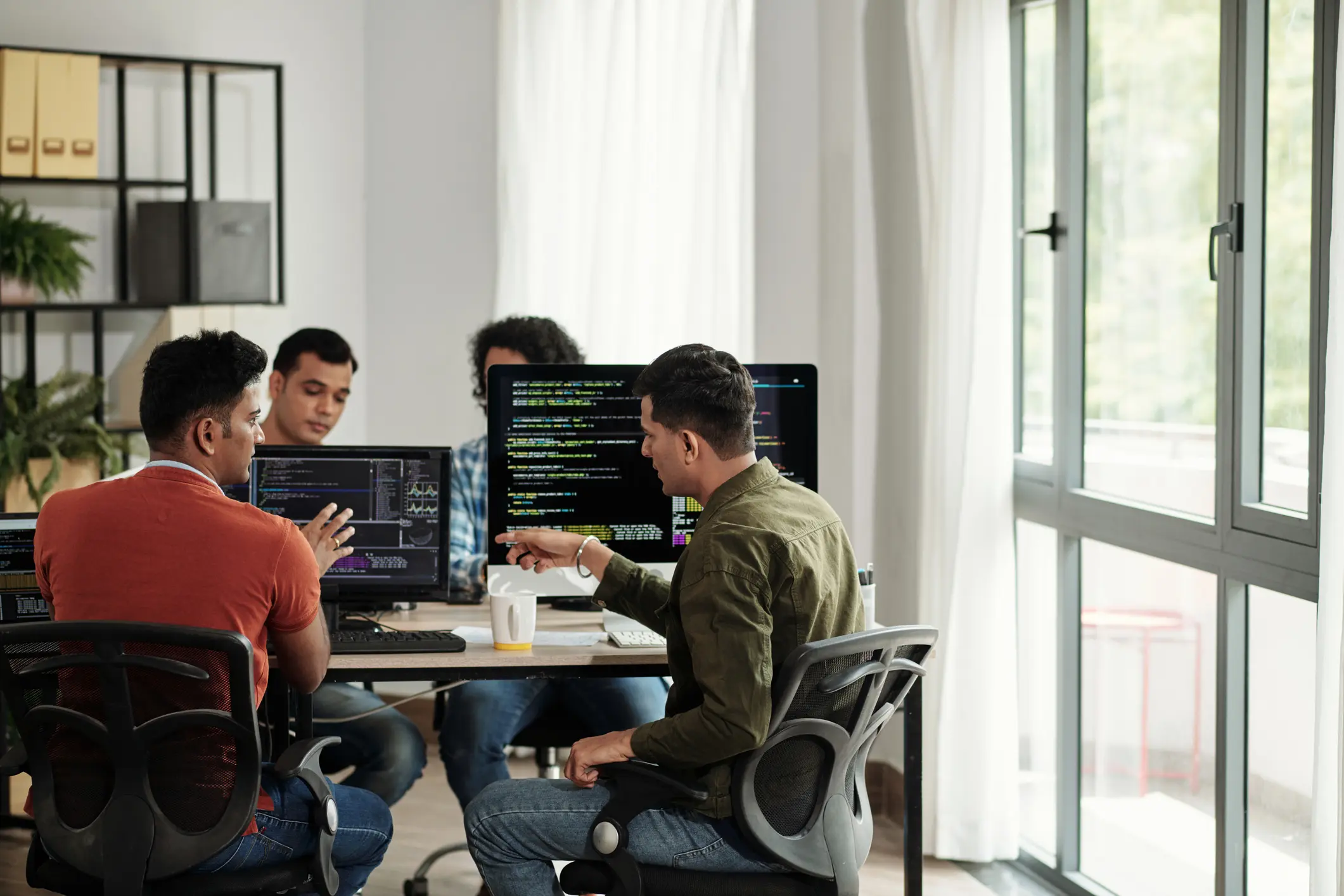 Four people collaborate around a table with laptops in a bright office.
