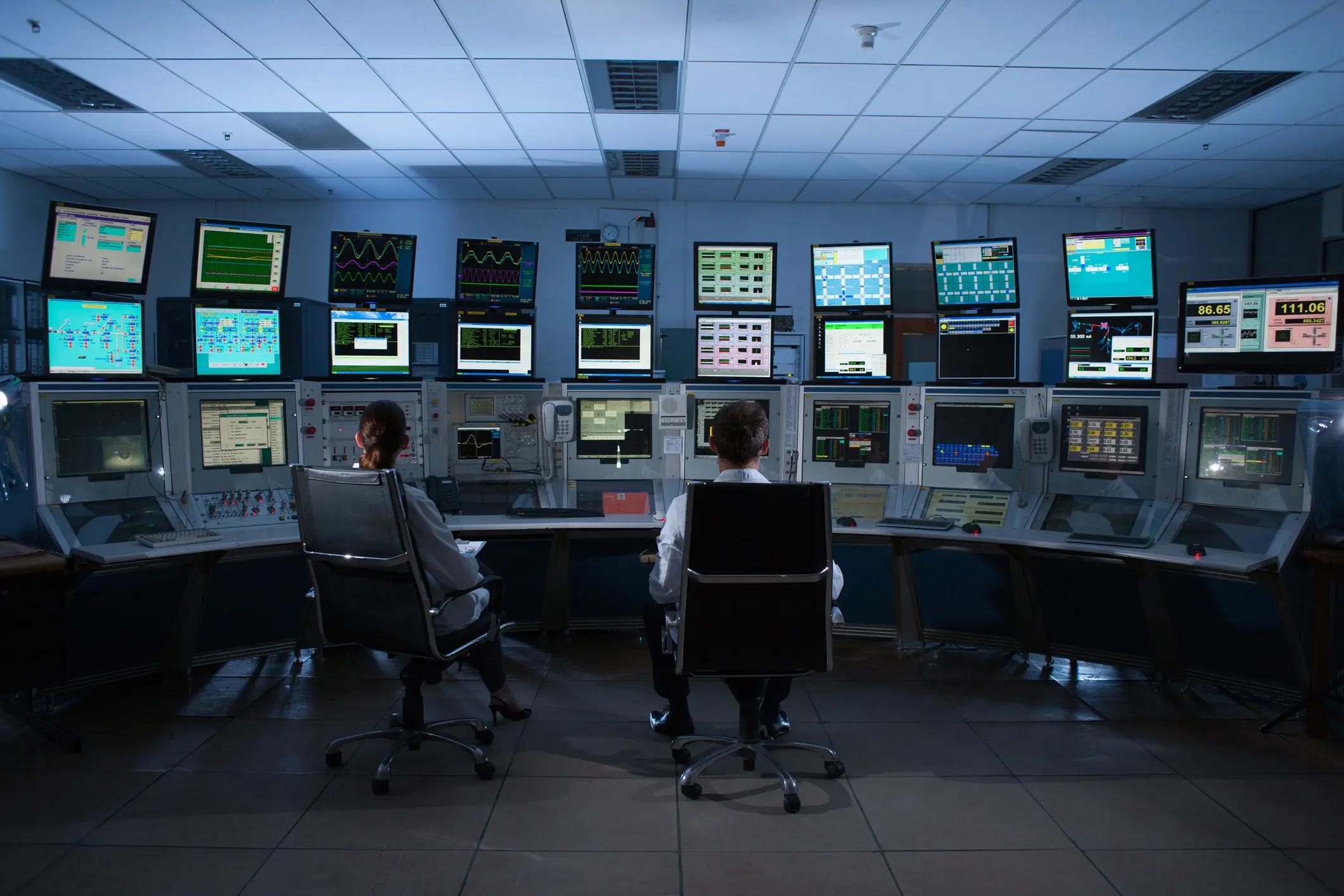 Two people monitor multiple screens in a control room filled with data and analytics.