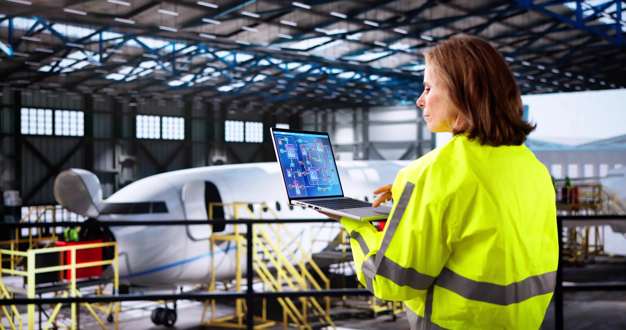A woman in high-visibility jacket using a laptop near an airplane inside a hangar.